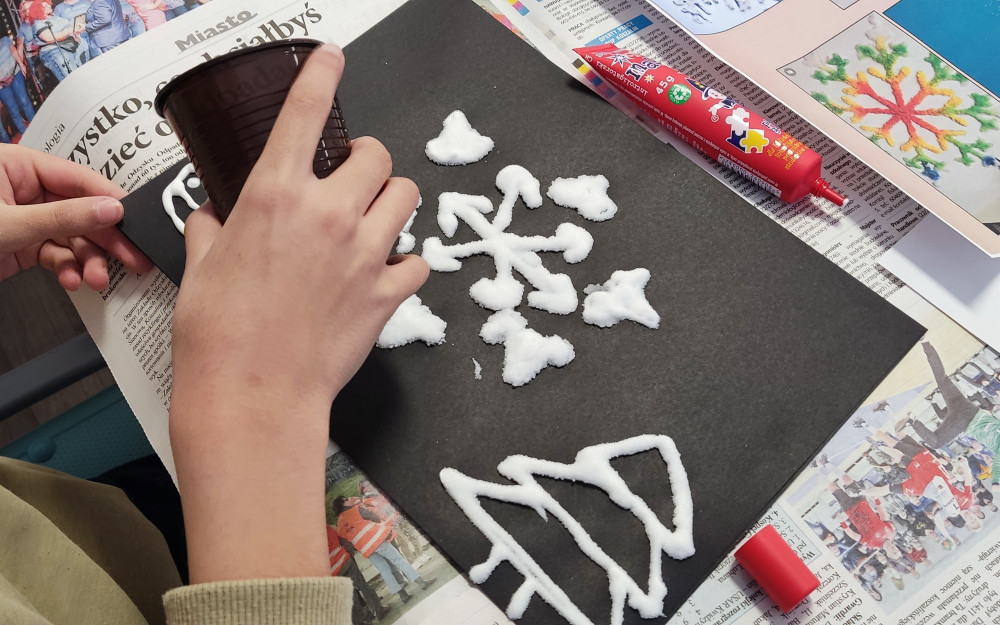 Na zdjęciu widoczne są dłonie dziecka wykonującego pracę plastyczną. Na czarnej kartce papieru znajduje się wzór śnieżynki oraz dwa trójkątne kształty przypominające choinki, wykonane z białej substancji (prawdopodobnie kleju i soli). Kartka leży na stole zabezpieczonym gazetami. Obok znajdują się materiały plastyczne, w tym czerwona tubka kleju. Dziecko trzyma w ręku plastikowy kubek.