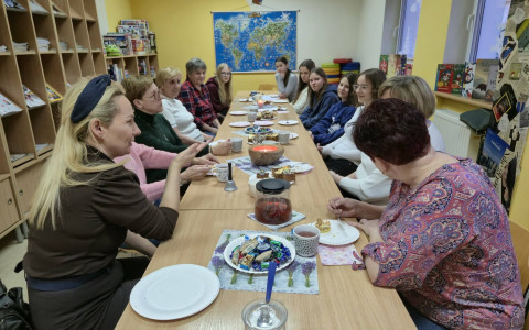 Na zdjęciu, przy długim stole siedzą COOLturalne 6 oraz uczennice SP nr 18 wraz z nauczycielką. Na stole słodki poczęstunek w postaci ciast i cukierków.