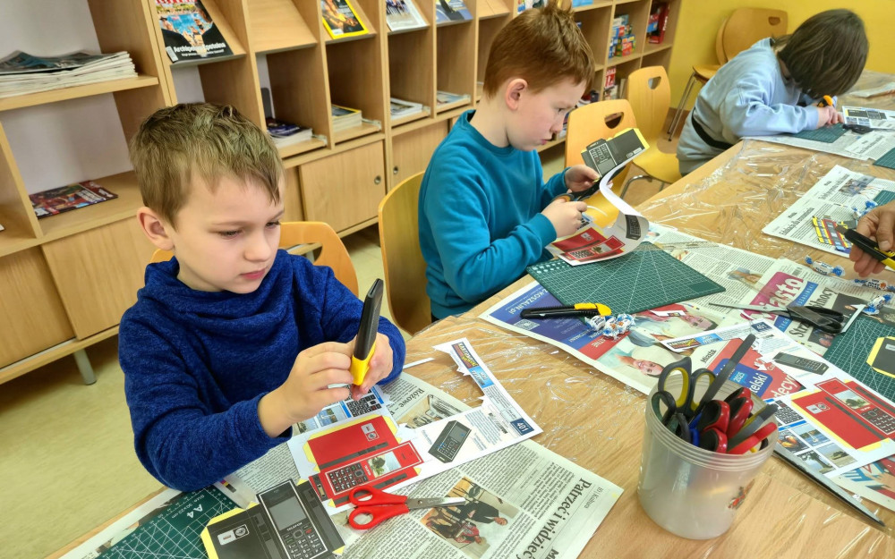 Na zdjęciu uczestnicy warsztatów modelarskich siedzą przy stole i wycinają z kartonu elementy telefonów komórkowych, które następnie będą sklejać.