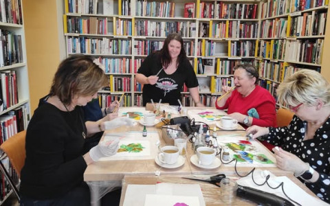 Uczestniczki spotkania wraz z bibliotekarką w czasie tworzenia prac, część pań polewa kartki tuszem, część rozdmuchuje tusz słomką.