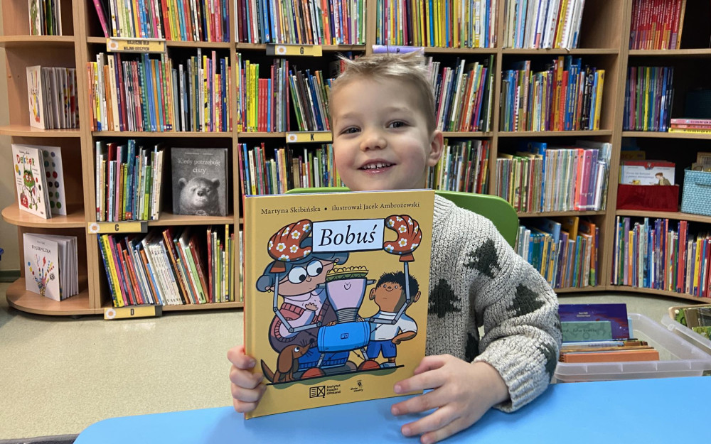 Chłopczyk siedzi na krześle, uśmiecha się do zdjęcia. w rękach trzyma książeczkę.