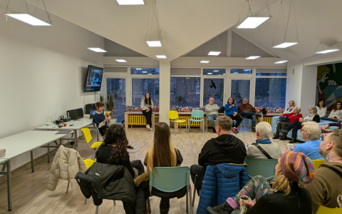 Jasne, nowoczesne wnętrze sali wykładowej. Grupa około 20-25 osób siedzi w dużym półkolu, przodem do prelegentki. Uczestnicy są w różnym wieku, siedzą na kolorowych krzesłach (żółtych i niebieskich) oraz błękitnych sofach. Po lewej stronie, przy ścianie z telewizorem, siedzi młoda kobieta z mikrofonem, która prowadzi spotkanie. W tle widać duże okna z widokiem na park o zmierzchu. Pomieszczenie jest oświetlone wieloma prostokątnymi lampami zwieszającymi się z sufitu.