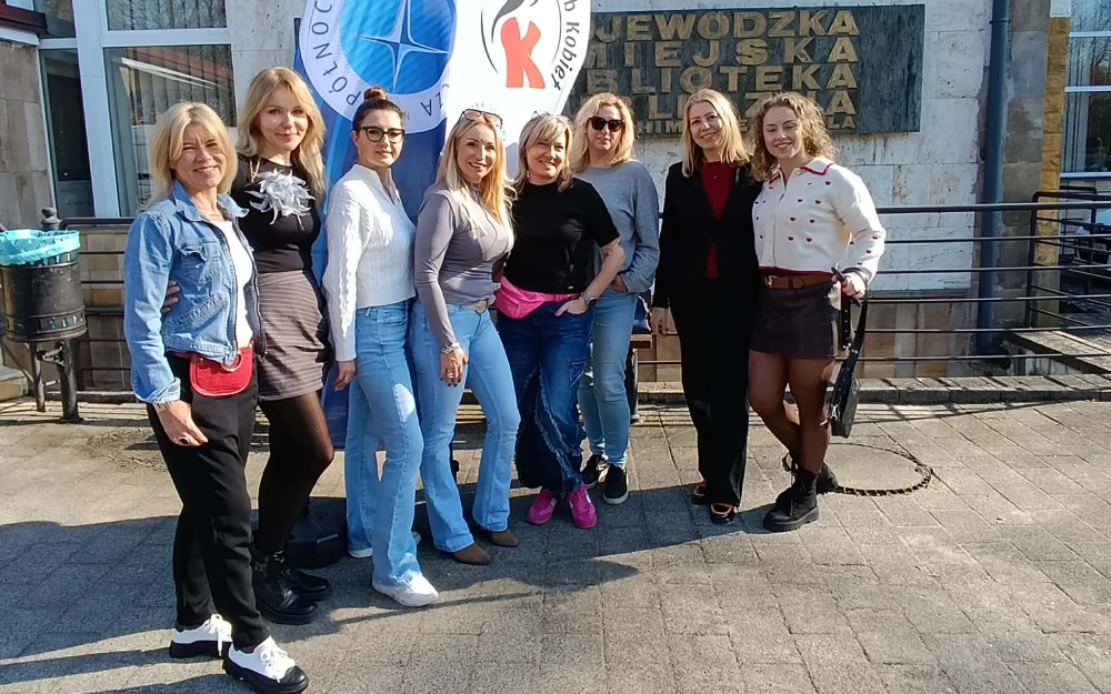 Zdjęcie przedstawia grupę ośmiu uśmiechniętych kobiet stojących na zewnątrz, przed budynkiem użyteczności publicznej w słoneczny dzień.