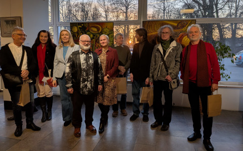 Zdjęcie grupowe dziesięciu osób pozujących w galerii na tle okien i obrazów. Wśród nich są artyści i goście w różnym wieku, ubrani w stroje casualowe i wizytowe. Kilka osób trzyma papierowe torby upominkowe. Za oknami widać zachodzące słońce i zarys drzew.