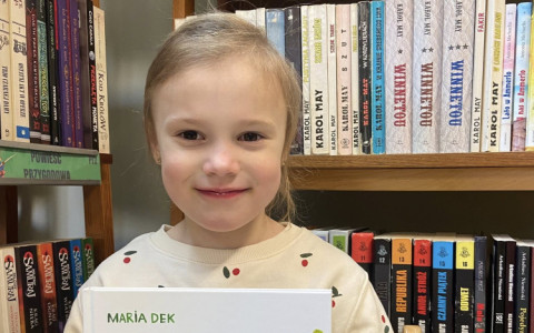 Dziewczynka prezentuje książkę i  uśmiecha się do aparatu.