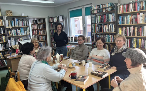Uczestniczki Dyskusyjnego Klubu Ksiązki siędzą przy Stole i rozmawiają o lekturze. Bibliotekarka stoi i uśmiecha isę do aparatu.