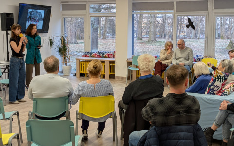 Na zdjęciu widać wnętrze jasnej sali z dużymi oknami, za którymi rozciąga się widok na drzewa bez liści. Po lewej stronie stoją dwie kobiety. Jedna z nich, młodsza, ma ciemne włosy, czarną koszulkę i szerokie jeansy; trzyma żółty mikrofon i przemawia. Obok niej stoi druga kobieta w niebieskiej kurtce, patrząc w stronę publiczności. Nad nimi, na ścianie, zawieszony jest telewizor wyświetlający zdjęcie architektury miejskiej.  Publiczność siedzi tyłem do obiektywu na kolorowych krzesłach (miętowych i żółtych) oraz na sofach w głębi. Grupa składa się z kilkunastu osób w różnym wieku. Wnętrze jest nowoczesne, z jasną podłogą, białymi ścianami i sufitowymi panelami LED. Na parapecie pod oknem leżą kolorowe poduszki w kwiatowe wzory.