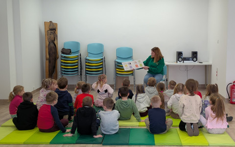 Zdjęcie przedstawia grupę dzieci siedzących na podłodze podczas czytania książki. W jasnej sali, na zielonych matach piankowych, siedzi tyłem około dwadzieścioro dzieci w wieku przedszkolnym. Przed nimi, na niebieskim krześle, siedzi nauczycielka w zielonej bluzie, która trzyma otwartą, ilustrowaną książkę i czyta ją grupie. W tle widać białe ściany, ułożone w stosy błękitne krzesła oraz drewnianą rzeźbę.