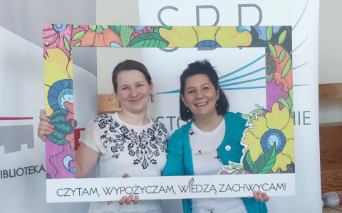 Dwie bibliotekarki pozują stojąc w kolorowej ramce z napisem "Czytam, wypożyczam, wiedzą zachwycam!"