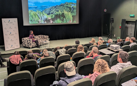 Widok z tyłu sali kinowej na scenę i widownię. Na scenie siedzi kobieta prowadząca spotkanie. Na ekranie widać zdjęcie panoramiczne górzystego krajobrazu pod zachmurzonym niebem. Widownia jest częściowo wypełniona osobami siedzącymi w ciemnozielonych fotelach kinowych. W tle po prawej stronie widać profesjonalny głośnik na statywie oraz drzwi wyjściowe.
