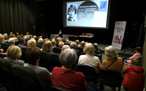 Szerokie ujęcie sali wykładowej z tyłu. Na ekranie widoczny slajd poświęcony Wisławie Szymborskiej (tytuł: „Dzieciństwo i młodość”), zawierający jej czarno-białe zdjęcie z młodości oraz informacje biograficzne. Na scenie, obok prelegentki, znajduje się stolik z ułożonymi książkami. Widownia, składająca się z kilkudziesięciu osób, siedzi w ciemnozielonych fotelach. fot. Adam Paczkowski