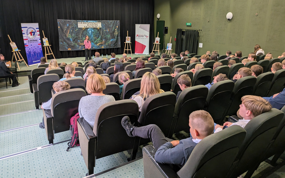 Szerokie ujęcie sali audytoryjnej z perspektywy ostatnich rzędów. Na środku oświetlonej sceny stoi młoda uczestniczka konkursu w różowej marynarce, która wygłasza swoją mowę. Przed nią znajduje się wypełniona widownia – widać plecy i profile osób siedzących w ciemnozielonych fotelach. Sala ma wysoki sufit z kasetonami i oliwkowe ściany.