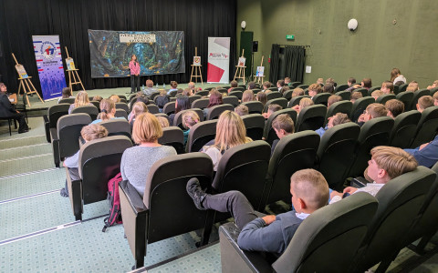 Szerokie ujęcie sali audytoryjnej z perspektywy ostatnich rzędów. Na środku oświetlonej sceny stoi młoda uczestniczka konkursu w różowej marynarce, która wygłasza swoją mowę. Przed nią znajduje się wypełniona widownia – widać plecy i profile osób siedzących w ciemnozielonych fotelach. Sala ma wysoki sufit z kasetonami i oliwkowe ściany.