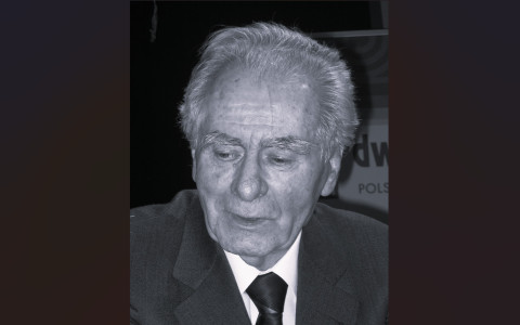 Czarno-biała fotografia portretowa przedstawiająca starszego mężczyznę w ujęciu od klatki piersiowej w górę. Mężczyzna jest zwrócony lekko w lewo, z wzrokiem skierowanym w dół. Ma siwe, zaczesane do tyłu włosy oraz krzaczaste, jasne brwi. Ubrany jest w ciemną marynarkę, jasną koszulę oraz ciemny krawat.  Tło jest ciemne i nieostre. Po prawej stronie, za ramieniem mężczyzny, widoczny jest fragment jasnej planszy z niepełnym napisem, w którym można rozpoznać litery „dw” oraz poniżej słowo „POLS”. Postać jest oświetlona z przodu, co wyraźnie podkreśla rysy twarzy. fot. Mariusz Kubik