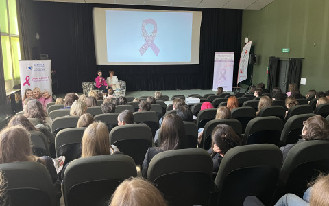 Zdjęcie przedstawia spotkanie edukacyjne odbywające się w nowoczesnej sali kinowej lub konferencyjnej. Tematyką wydarzenia jest profilaktyka raka piersi, na co wskazują różowe symbole i napisy w języku polskim.  Szczegóły obrazu  Pierwszy plan: Widzimy tył głów osób siedzących w rzędach ciemnych, tapicerowanych foteli. Publiczność składa się w przeważającej mierze z kobiet.  Drugi plan: Na niewielkiej scenie, po lewej stronie, siedzą dwie kobiety. Jedna z nich ma na sobie jaskraworóżową marynarkę i trzyma mikrofon, co sugeruje, że prowadzi prelekcję lub dyskusję. Obok nich stoi baner reklamowy z napisem „Szpital Wojewódzki w Koszalinie” oraz hasłem „Dbam o piersi! Profilaktyka raka piersi”.  Tło: W centralnym punkcie znajduje się duży ekran projekcyjny, na którym wyświetlona jest grafika przedstawiająca dużą różową wstążkę (symbol walki z rakiem piersi) z napisem „PIER(W)SI NA BADANIA”. Po prawej stronie ekranu stoi kolejny pionowy baner z tym samym hasłem.  Otoczenie: Sala ma ciemnozielone ściany i czarne kotary po bokach ekranu. Oświetlenie jest stonowane, punktowe, umieszczone w podwieszanym suficie. Po prawej stronie widoczne są czarne drzwi wyjściowe z zielonym znakiem ewakuacyjnym powyżej.  Kontekst Zdjęcie dokumentuje wydarzenie prozdrowotne skierowane do lokalnej społeczności, mające na celu podniesienie świadomości na temat regularnych badań profilaktycznych.