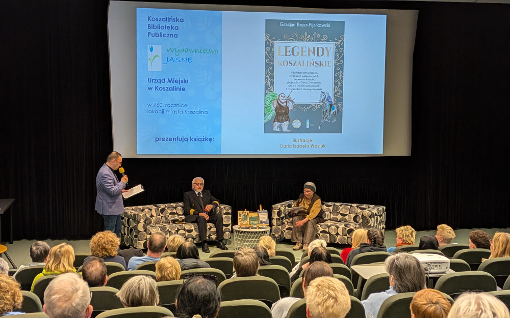 Widok na scenę w sali kinowej podczas spotkania autorskiego. Na dużym ekranie wyświetlona jest okładka książki „Legendy Koszalińskie” Gracjana Bojara-Fijałkowskiego oraz logotypy organizatorów: Koszalińskiej Biblioteki Publicznej, Wydawnictwa Jasne i Urzędu Miejskiego w Koszalinie. Pod ekranem, na wzorzystych fotelach, siedzą dwie osoby: starszy mężczyzna w ciemnym mundurze marynarskim oraz starsza kobieta w brązowej kamizelce i opasce na głowie. Po lewej stronie stoi Dyrektor KBP z mikrofonem. W dolnej części zdjęcia widać głowy licznie zgromadzonej publiczności siedzącej w rzędach.