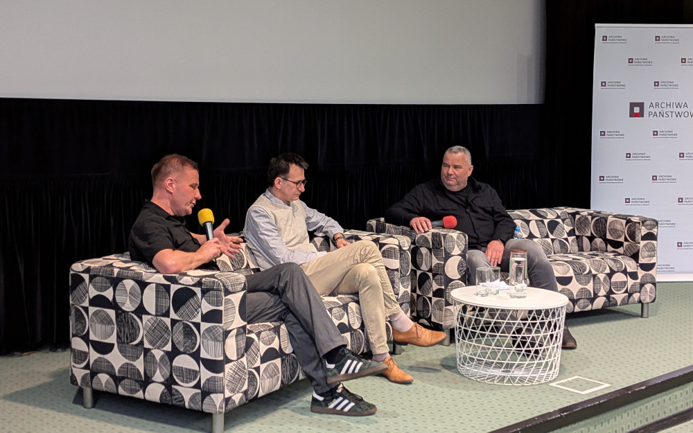 Zbliżenie na scenę, na której trwa panel dyskusyjny. Trzech mężczyzn siedzi na dwóch kanapach o geometrycznym, czarno-białym wzorze. Mężczyzna po lewej, ubrany w czarną koszulę, mówi do mikrofonu, gestykulując. W środku siedzi mężczyzna w jasnym swetrze i okularach (prowadzący z pierwszego zdjęcia), a po prawej mężczyzna w ciemnej koszuli. Między kanapami stoi biały, ażurowy stolik, na którym znajdują się szklanki i karafka z wodą. W tle widoczny jest baner „ARCHIWA PAŃSTWOWE”.