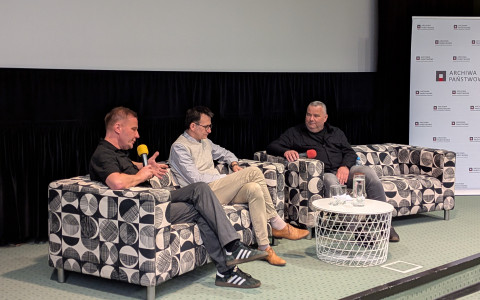 Zbliżenie na scenę, na której trwa panel dyskusyjny. Trzech mężczyzn siedzi na dwóch kanapach o geometrycznym, czarno-białym wzorze. Mężczyzna po lewej, ubrany w czarną koszulę, mówi do mikrofonu, gestykulując. W środku siedzi mężczyzna w jasnym swetrze i okularach (prowadzący z pierwszego zdjęcia), a po prawej mężczyzna w ciemnej koszuli. Między kanapami stoi biały, ażurowy stolik, na którym znajdują się szklanki i karafka z wodą. W tle widoczny jest baner „ARCHIWA PAŃSTWOWE”.