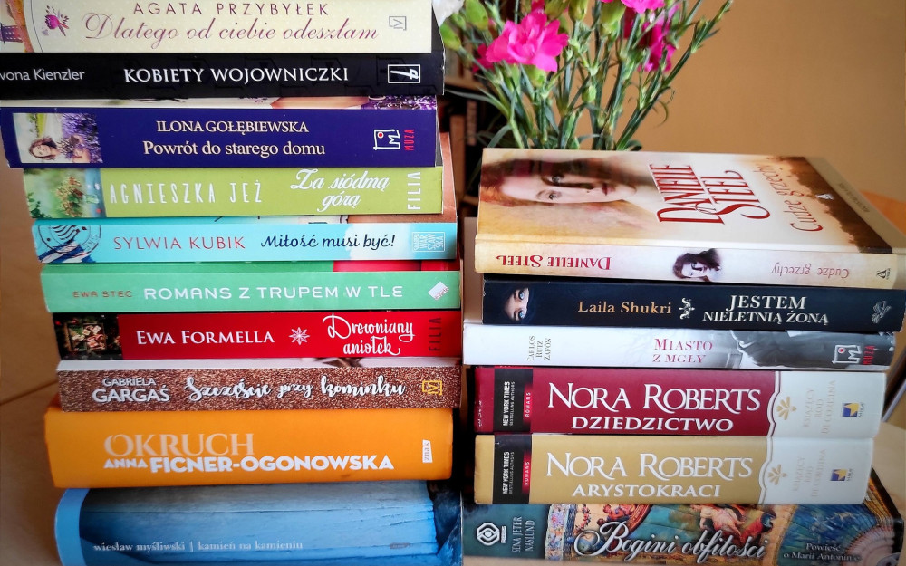 Dwa stosy książek różnych autorów ustawione na stole, obok wazon z różowymi kwiatami. W tle rozmyty regał z książkami.