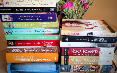 Dwa stosy książek różnych autorów ustawione na stole, obok wazon z różowymi kwiatami. W tle rozmyty regał z książkami.