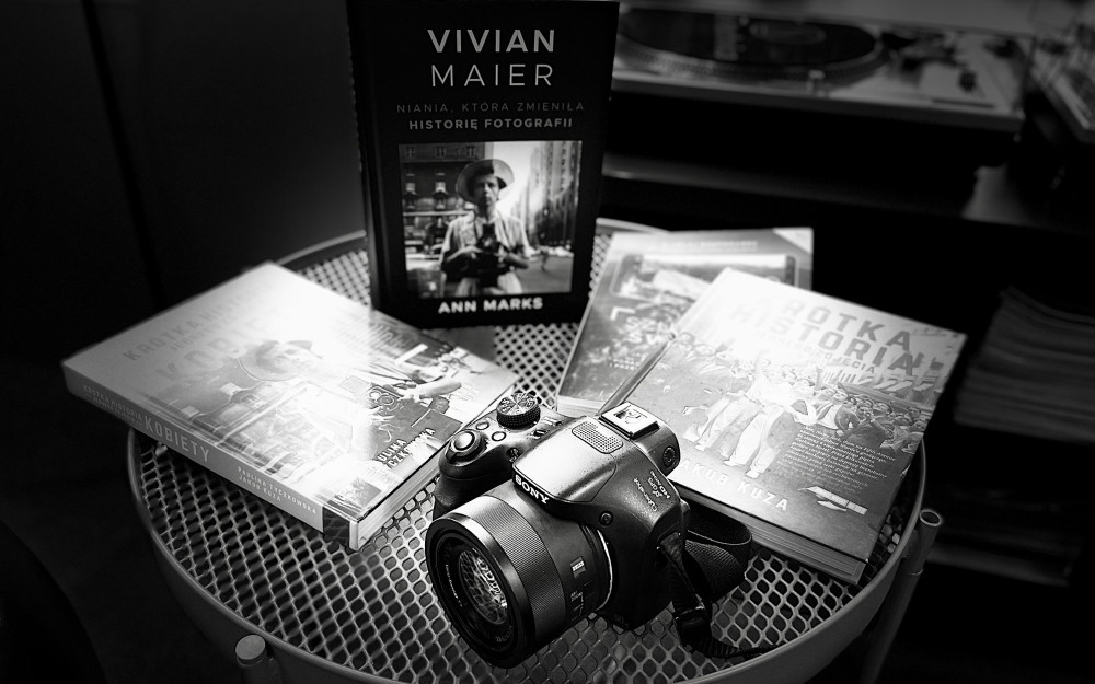 Czarno-białe, artystyczne zdjęcie przedstawiające aparat fotograficzny oraz kilka książek ułożonych na okrągłym stoliku z metalowej siatki. Kompozycja ma charakter martwej natury i nawiązuje do tematyki historii fotografii.