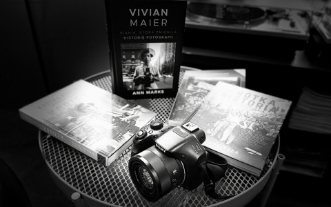 Czarno-białe, artystyczne zdjęcie przedstawiające aparat fotograficzny oraz kilka książek ułożonych na okrągłym stoliku z metalowej siatki. Kompozycja ma charakter martwej natury i nawiązuje do tematyki historii fotografii.