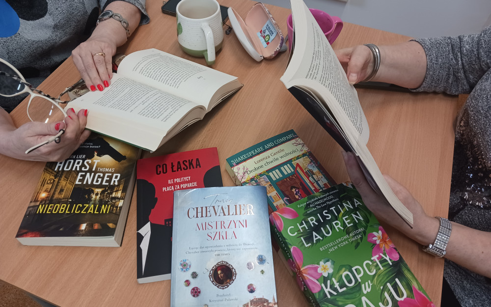 Dwie panie trzymają w rękach książki, na stole leżą książki, dwa kubki i okulary do czytania