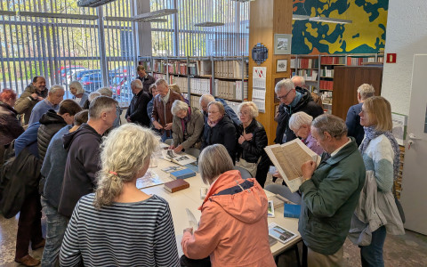 Wnętrze biblioteki. Grupa seniorów tłoczy się wokół dużego, jasnego stołu, na którym wyłożone są stare dokumenty, księgi i albumy. Uczestnicy z zainteresowaniem przeglądają materiały. W tle widać regały z segregatorami oraz dużą, kolorową abstrakcyjną grafikę na ścianie.