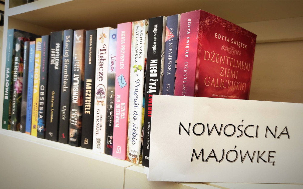 Na półce bibliotecznej stoją nowe książki dla dorosłych czytelników. Obok nich znajduje się kartka z napisem „Nowości na majówkę”.