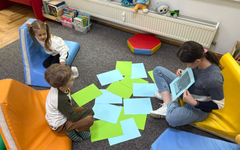 Dzieci wraz z bibliotekarką siedzą na dywanie, bibliotekarka pokazuje im kolorowe obrazki