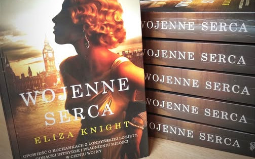 Ksiażki ułożone w stos, jedna stoi przodem, książki pt "Wojenne serca" Elizy Knight, w tle fioletowe bzy.