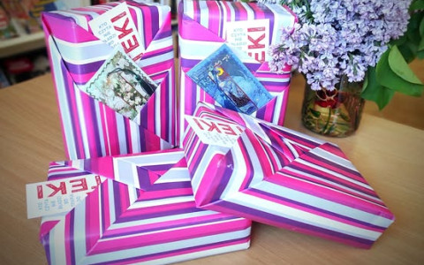 4 zapakowane w pasiasty różowo fioletowy papier niespodzianki książkowe. w tle fioletowe bzy i regał z książkami.