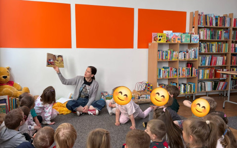 Bibliotekarka czyta dzieciom książkę, dzieci siędza na szarym dywanie