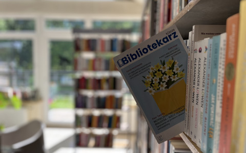 Na pierwszym planie widoczny jest regał biblioteczny z książkami. Pomiędzy książkami umieszczony jest egzemplarz czasopisma „Bibliotekarz” z okładką przedstawiającą ilustrację żółtych kwiatów wyrastających z książki. W tle, lekko rozmytym, znajduje się jasne wnętrze biblioteki z kolejnymi regałami i dużymi oknami wpuszczającymi naturalne światło.