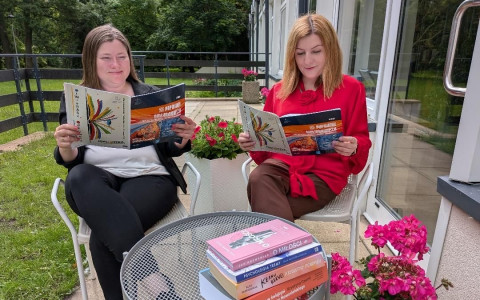 Na tarasie przed budynkiem dwie kobiety siedzą przy okrągłym stoliku i czytają czasopismo. Na stoliku leży stos kilku książek. Wokół znajdują się donice z różowymi kwiatami. W tle widoczny jest trawnik, drzewa oraz przeszklona ściana budynku.