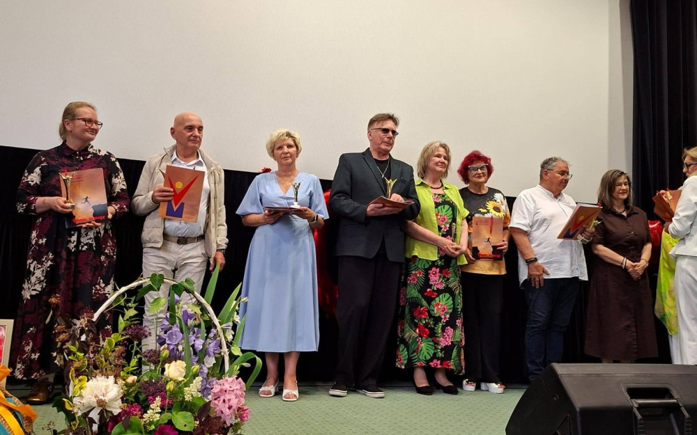 Na scenie sali widowiskowej stoi grupa kobiet i mężczyzn uczestniczących w jubileuszu 10-lecia Stowarzyszenia VivaMY w Koszalinie. Kilka osób trzyma pamiątkowe statuetki i dyplomy. W tle znajduje się duży ekran projekcyjny, a przed sceną widoczna jest kompozycja kwiatowa.
