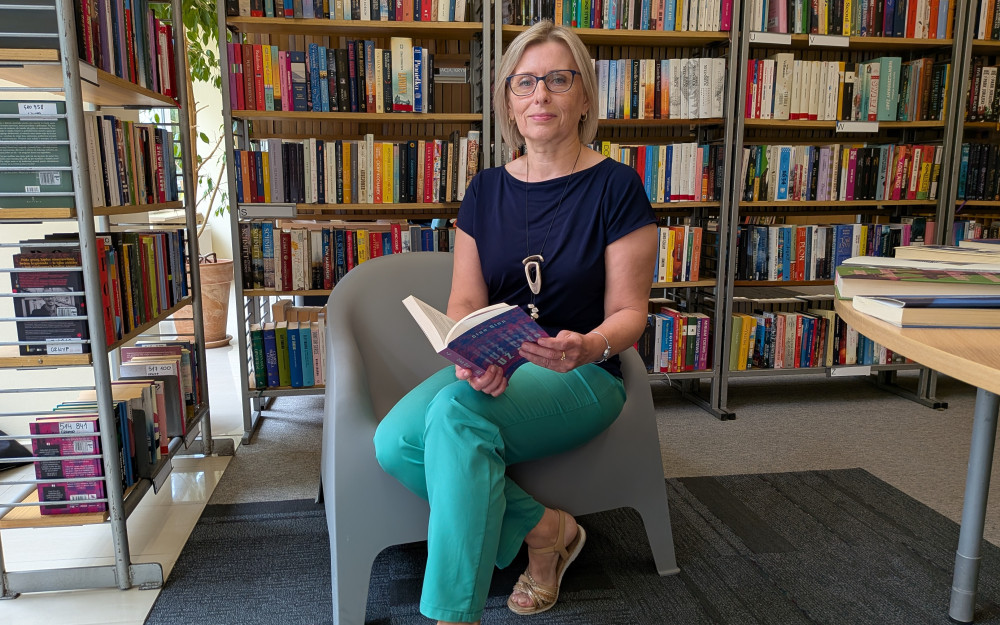 Na zdjęciu widać Bogusławę Kałużną (wicedyrektor KBP) siedzącą w szarym fotelu pomiędzy regałami z książkami w bibliotece. Trzyma w dłoniach otwartą książkę i patrzy w obiektyw z delikatnym uśmiechem. Ubrana jest w granatową bluzkę, turkusowe spodnie i jasne sandały, ma okulary oraz długi naszyjnik. W tle znajdują się wysokie regały wypełnione książkami, a po prawej stronie widoczny jest fragment stołu z ułożonymi publikacjami. Wnętrze jest dobrze oświetlone światłem dzienny