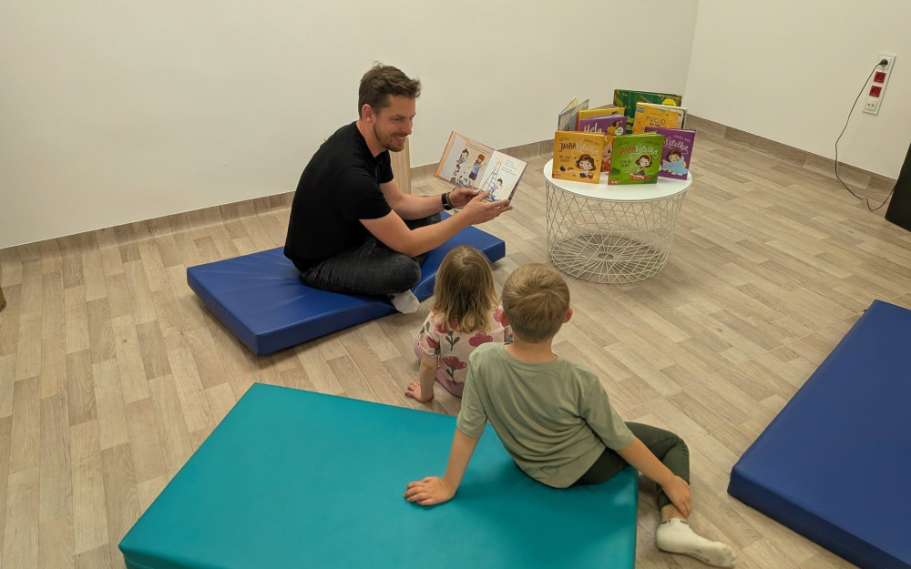 Mężczyzna siedzi na materacu i czyta książkę dwojgu małych dzieci siedzących przed nim. Obok znajduje się niski stolik z książkami dla najmłodszych. Spotkanie odbywa się w sali z materacami do zabawy.