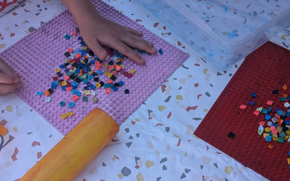 Na stole przykrytym jasnym obrusem dziecko układa kolorowe elementy LEGO DOTS na różowej płytce konstrukcyjnej. Obok leży wałek do nakładania farby oraz pojemnik z materiałami do warsztatów.