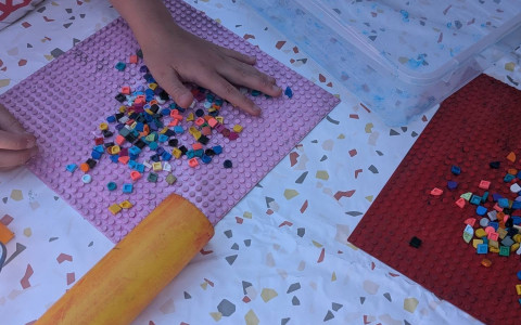 Na stole przykrytym jasnym obrusem dziecko układa kolorowe elementy LEGO DOTS na różowej płytce konstrukcyjnej. Obok leży wałek do nakładania farby oraz pojemnik z materiałami do warsztatów.
