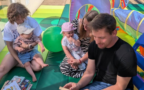 Na kolorowych matach przed budynkiem biblioteki siedzą dwie osoby dorosłe z dwójką małych dzieci. Jeden z dorosłych czyta książkę, obok leży stos książek dla najmłodszych. W tle znajduje się kolorowy tunel i namiot do zabawy oraz piłka rehabilitacyjna.