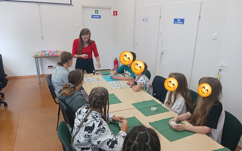 Dzieci siedzą przy stolikach, prowadząca warsztaty stoi  i pokazuje dzieciom butelki z olejkami