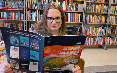 Kobieta w okularach siedzi w bibliotece, przed regałami wypełnionymi książkami. Trzyma otwarte czasopismo „Poradnik Bibliotekarza” i patrzy w stronę aparatu.