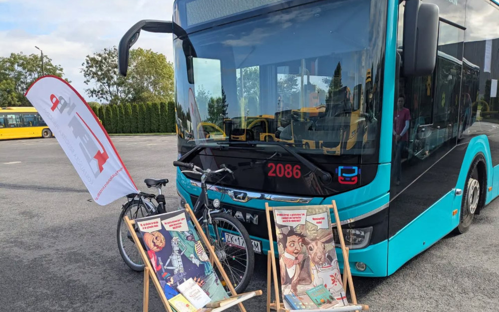 Turkusowy autobus MZK z biblioteczną aranżacją przed wejściem. Obok stoją dwa leżaki z okładkami książek, rower i flaga Koszalińskiej Biblioteki Publicznej.