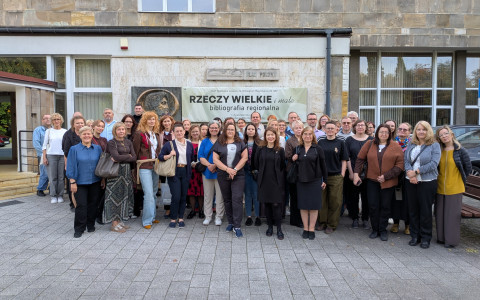 Pamiątkowe zdjęcie grupowe uczestników konferencji stojących na zewnątrz przed wejściem do budynku biblioteki. W tle widać baner z nazwą wydarzenia „Rzeczy wielkie i małe – bibliografia regionalna”.