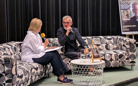 Zbliżenie na scenę z ciemną kurtyną. Wojciech Tochman w okularach i ciemnym stroju mówi do czerwonego mikrofonu, siedząc na wzorzystej kanapie. Obok na sąsiedniej kanapie siedzi prowadząca w białej koszuli z żółtym mikrofonem. Przed nimi stoi mały, ażurowy stolik z książką na sztaludze i szklankami wody, a obok plakat autorski.