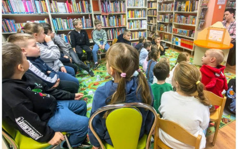 Uczniowie klasy 1b siedzą wśród regałów z książkami i słuchają opowiadania prezentowanego w teatrzyku kamishibai.