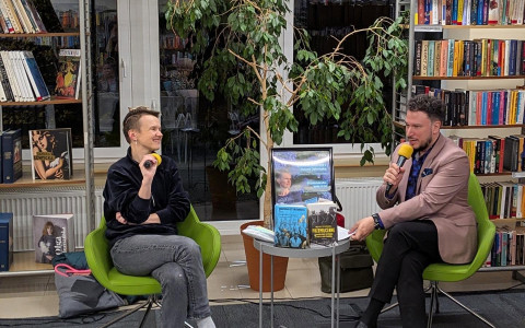 Rozmowa podczas spotkania autorskiego w bibliotece. Dwie osoby siedzą naprzeciwko siebie na zielonych fotelach i trzymają mikrofony z żółtymi osłonami. Na stoliku między nimi leżą książki. W tle widoczne są regały biblioteczne i rośliny.