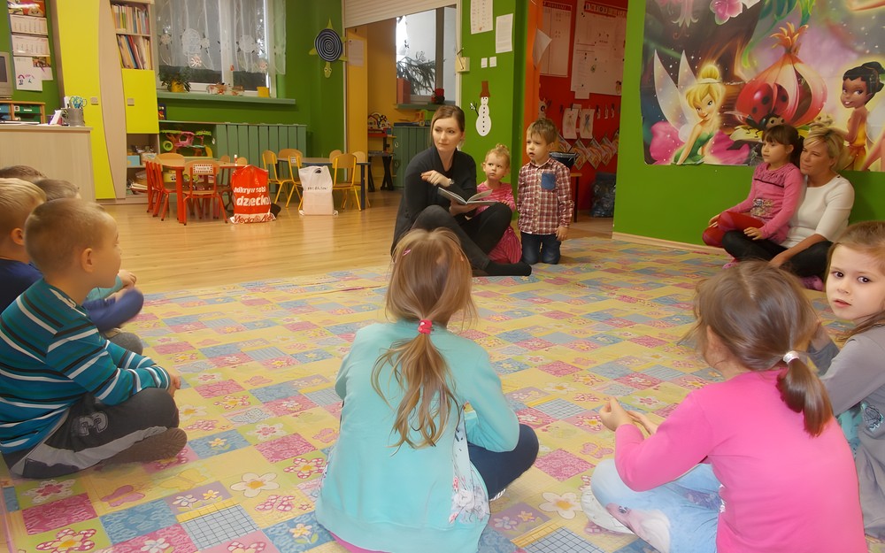 Dzieci siedzą na dywanie i słuchają bibliotekarki, która czyta książkę