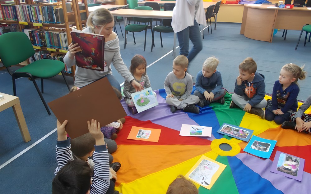 Dzieci siedzą na chuście i oglądają ilustracje pokazywane przez bibliotekarkę